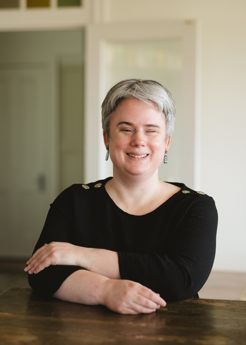 Lauren Hayes laughing, seated at a wooden table with arms folded
